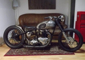 Moto Triumph bobber color metal sobre una alfombra persa en una sala con sofá y lampara